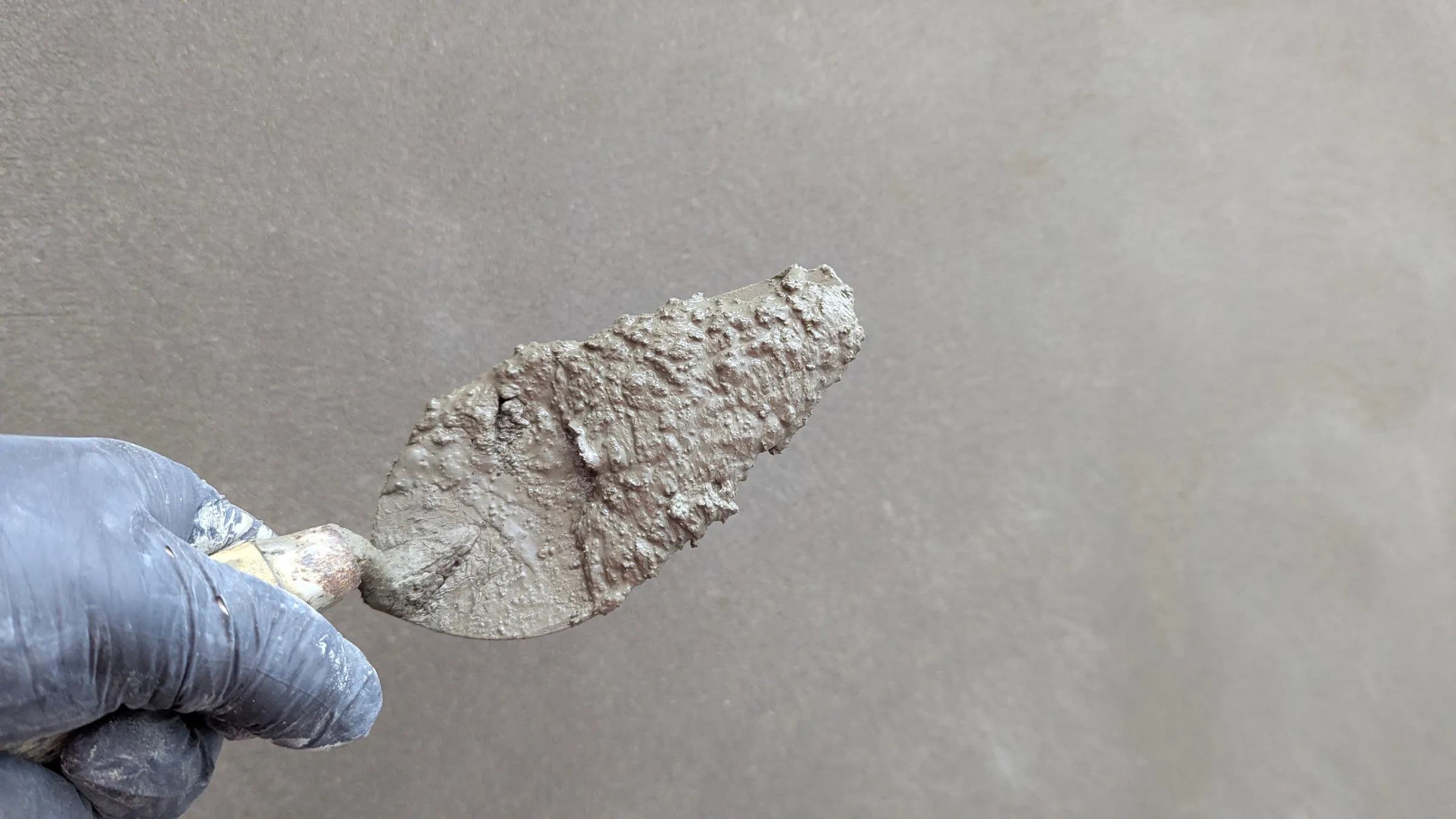 Stock photo: a gloved hand holding a pointed trowel loaded with wet cement plaster in front of a smooth gray wall.